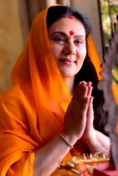 a woman in an orange sari sitting at a table with her hands clasped up