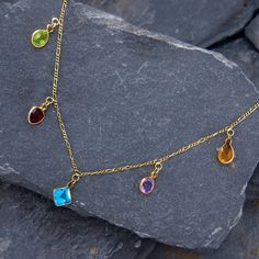 This bejeweled station necklace features a pinch bezel-set oval cut peridot, heart cut garnet, square cut blue topaz, round cut amethyst, and pear cut citrine that dangle from an 18 inch long baby figaro chain. The necklace is crafted in 14k yellow gold and finished with a spring ring clasp. 14k Gold Jewelry With Gemstone Accents For May Birthstone, Gold Multi-stone Fine Jewelry Gemstones, Gold Multi-stone Gemstones In Fine Jewelry Style, Luxury Drop Multi-stone Jewelry, 14k Gold Teardrop Jewelry With Gemstone Accents, Elegant Multicolor Gemstones In Bezel Setting, Elegant Multicolor Gemstones With Bezel Setting, Gold Multi-stone Jewelry For May Birthstone, Elegant Multi-stone Drop Necklaces