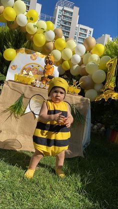 a baby dressed as a bee holding a tennis racquet in front of balloons