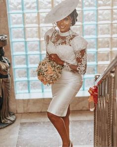 a woman in a white dress and hat holding a bouquet standing next to a stair case