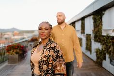 a man and woman are standing on a balcony with flowers in the foreground, looking at the camera