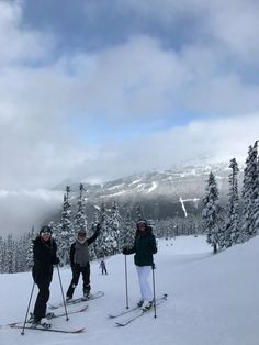 three people on skis standing in the snow with trees and mountains in the background