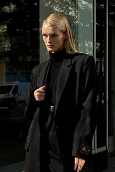 a woman standing on the sidewalk in front of a building wearing a black coat and pants