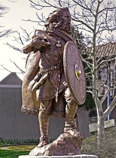 a statue of a man in armor holding a shield and standing next to a tree