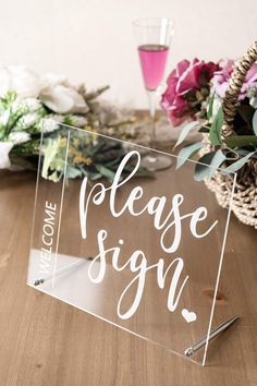 a clear acrylic sign sitting on top of a wooden table next to flowers