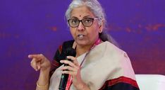 an old woman with glasses speaking into a microphone in front of a purple wall and white chair