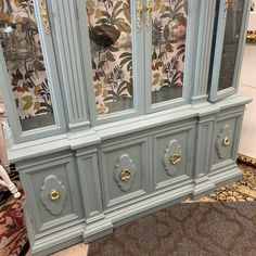 a blue china cabinet with floral wallpaper on the back and gold handles, in a living room