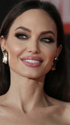 a woman with long dark hair wearing large hoop earrings and black eyeshadow, smiling at the camera