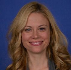 a woman with blonde hair smiling at the camera and wearing a gray jacket, against a blue background