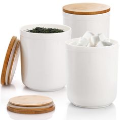 three white containers with wooden lids and some sugar cubes on the top one is filled with green tea