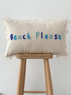 a white pillow with the word beach please embroidered on it sitting on a wooden stool