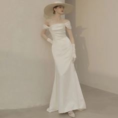 a woman in a white dress and hat standing next to a wall with her hands on her hips