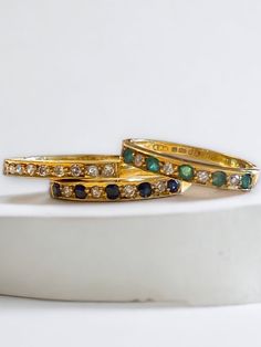 two gold rings with emeralds and white diamonds on a marble stand against a white background