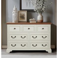 a white dresser with drawers and vases on top in front of a framed photograph