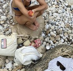 a woman is sitting on the rocks with an apple in her hand and other items around her