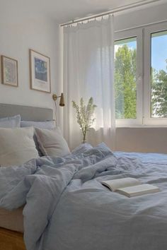 an unmade bed with blue sheets and pillows in front of two large windows that look out onto the trees outside