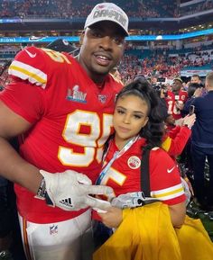 a man standing next to a woman in a football uniform on the sidelines at a stadium