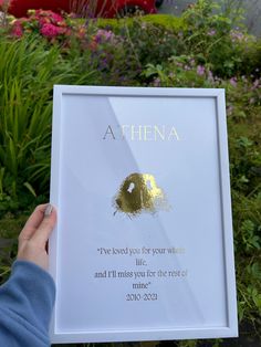 a person holding up a white framed sign in front of some flowers and plants with the words athena written on it