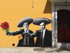 two men wearing sombreros and holding flowers in front of a wall with a painting on it