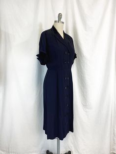 "Vintage 1950s dress featuring; ~ a v neck with notched collar short sleeves with winged cuff lightly padded shoulders collar and cuff are trimmed in velvet has two rows of buttons all the way down the front string belt loops at the waist but no belt skirt is fairly straight and falls to below the knee ~ of a mid weight faille fabric in a deep navy blue ~ labeled \"Make mine a McKettrick\" this has no size label, measurements when laid out flat are: 23 inches from pit to pit 10 inch sleeve 2 1/2 Vintage Navy Dress For Formal Occasions, 1950s Style Vintage Short Sleeve Dress For Work, Vintage Dress With Covered Buttons And Short Sleeves, Formal Vintage Dress With Short Sleeves And Buttons, Navy Vintage Style Formal Dress, Retro Vintage Dress With Short Sleeves For Formal Occasions, 1950s Style Short Sleeve Vintage Dress For Formal Occasions, Vintage Short Sleeve Dress For Work, Vintage Short Sleeve Dress With Covered Buttons