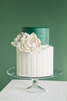 a white and green wedding cake with flowers on top is sitting on a glass platter