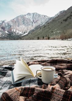 an open book and coffee cup on a blanket by the water with mountains in the background