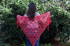 a woman wearing a red crocheted shawl standing in front of some bushes