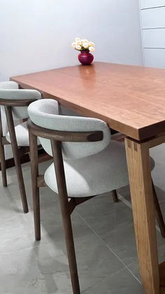 An image of a dining area featuring a sleek, rectangular wooden table with a natural finish, accompanied by modern chairs with light gray upholstery and dark wooden frames. The chairs have a curved backrest for added comfort. On the table sits a striking purple vase holding a bouquet of cream-colored flowers, adding a pop of color to the scene. The setting is minimalistic, with a gray wall and light gray tiled flooring, evoking a contemporary and clean dining space. Urban Home Decor, Home Decor Scandinavian, Purple Vase, Cream Flowers, Scandi Style