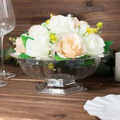 a glass bowl filled with flowers on top of a wooden table next to wine glasses