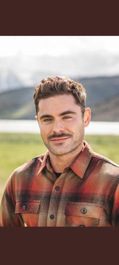 a man with a mustache standing in front of a field and mountains, wearing a flannel shirt