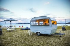 an old camper trailer is parked on the grass by the water with tables and chairs around it