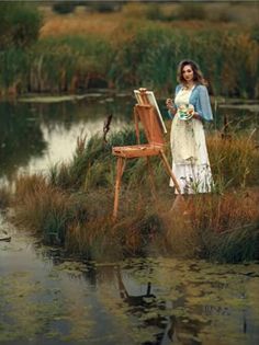 a woman standing next to an easel in the grass near a pond with a bird on it