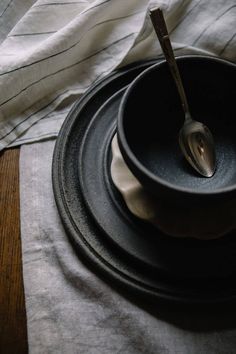 a black bowl and spoon sitting on top of a white cloth covered placemat next to a wooden table