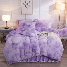 a bed with purple comforter and pillows in a room next to a lamp on a table