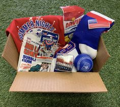 an open cardboard box with sports items in it on the grass, including t - shirts and gloves