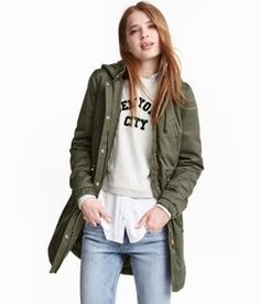 a young woman is standing in front of a white background wearing an army green jacket and jeans