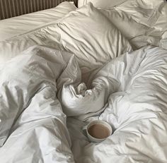 an unmade bed with white sheets and pillows on top of it, next to a cup of coffee