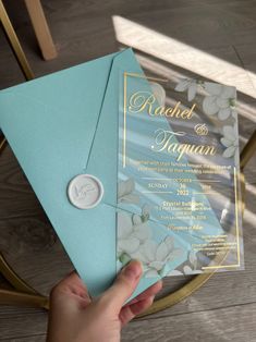 a person holding up a blue and gold wedding card with white flowers on the inside