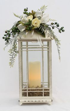 a white lantern with flowers and greenery on the top is lit by a candle