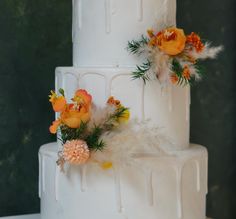 a three tiered white cake with orange flowers and feathers on the top, sitting on a table