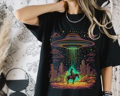 Step into the unknown with our "Cowboy in the Desert" UFO T-Shirt! Featuring a neon, retro-inspired design of a cowboy riding through the desert, suddenly illuminated by a mysterious UFO, this shirt brings a one-of-a-kind blend of western vibes and sci-fi intrigue to your wardrobe. Why You'll Love It: Bold Design: The unique combination of cowboy culture and extraterrestrial adventure makes this shirt a must-have for anyone looking to stand out. Material: 100% ring-spun cotton for a soft and dur Alien Desert, Desert Cowboy, Cowboy Culture, Neon Retro, Western Vibes, Desert Design, Cowboy Shirt, Alien Abduction, Cowboys Shirt