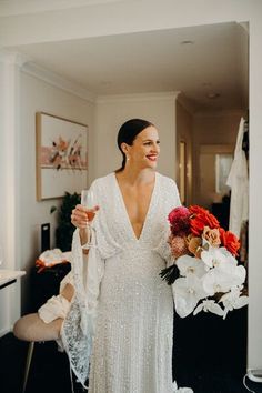 a woman in a white dress holding a glass of wine and standing next to a flower bouquet