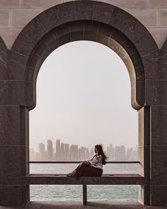 a woman sitting on top of a bench next to the ocean under an arched window