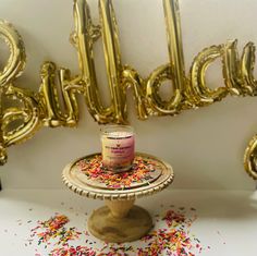 a birthday cake with sprinkles and a candle on a pedestal in front of balloons