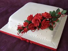 a white cake with red roses on it sitting on a purple cloth covered tablecloth