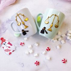 two coffee mugs with gold earrings on them next to marshmallows and candies