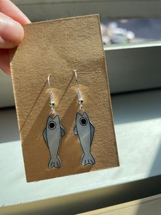 a pair of fish shaped earrings sitting on top of a piece of cardboard next to a window