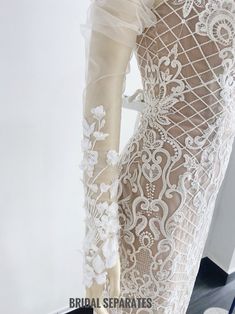 the back of a wedding dress with white lace and flowers on it, in front of a mannequin