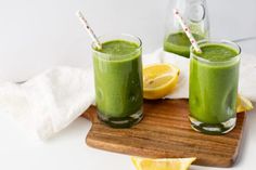 two glasses filled with green smoothie on top of a cutting board next to sliced lemons