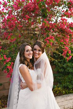 two women in wedding gowns standing next to each other with pink flowers behind them